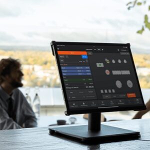 Monitor displaying a data dashboard with charts and tables on a stand at a table, a blurred person in the background.