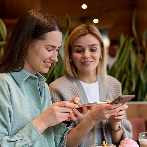 Two women sit together in a cafe, smiling as they look at a shared smartphone screen.