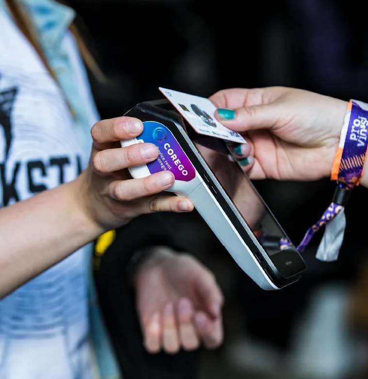 Person swipes a card over a handheld card reader; a second person holds the reader and has a purple wristband nearby