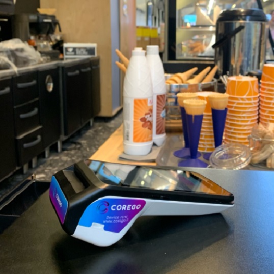 Card payment terminal (CoreGO) on a cafe counter, with orange cups and bottles in the background.
