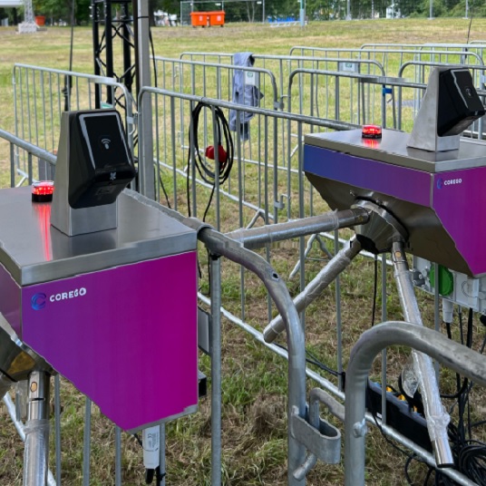 Outdoor barrier gate system with two purple Corego access terminals and red beacon lights on metal rails at an event entrance.