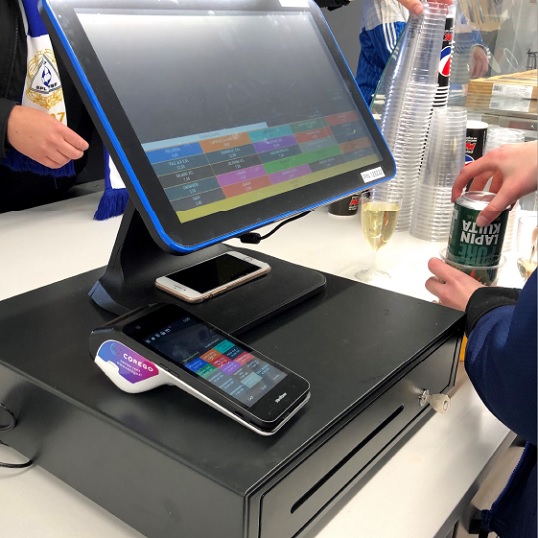 POS touchscreen checkout with card reader and smartphone on the counter; a customer with a drink at the register