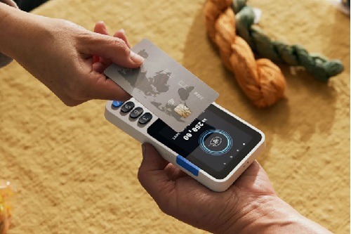 Two hands exchange a credit card over a small handheld payment terminal on a yellow surface.