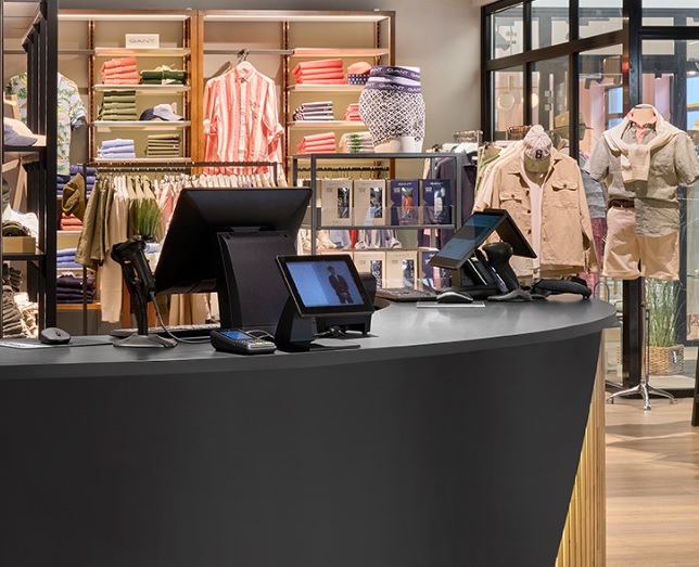 Checkout counter in a clothing store with card readers and screens, shelves of folded clothes and hanging garments visible in the background.